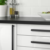 Modern kitchen with white cabinets and black handles. Countertop holds a bowl of salad and oil dispensers. Plants add greenery.