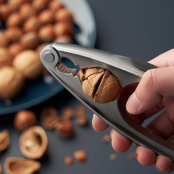 Hand holding nutcracker squeezing nut; nuts scattered around, blue plate in background.