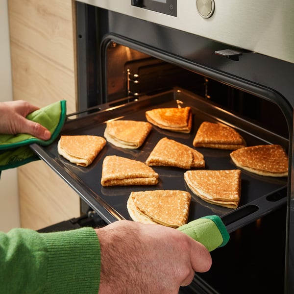 Person pulls out tray of golden-brown PANNKAKOR pancakes from oven. Green oven mitts used.