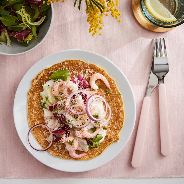 An overhead view of a savory pancake topped with shrimp, red onions, and greens on a white plate next to pink cutlery and a bowl of salad.