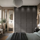 Large, dark grey PAX wardrobe with four doors, positioned beside a mirror. It includes a bed with blue cover and a nightstand with a lamp.