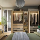 Modern bedroom with PAX wardrobe, featuring hanging clothes, shelves, and internal lighting.