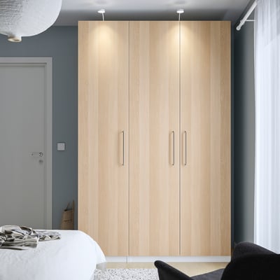 Minimalist bedroom with a tall light wood PAX wardrobe, white bed, and black chair against a dark wall.