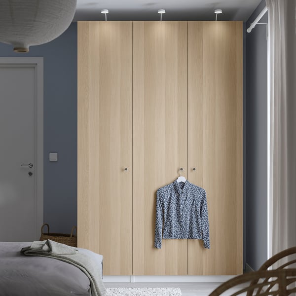 Minimalist bedroom with PAX wardrobe, patterned shirt, bed, and wicker chair by blue door.