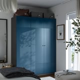 Blue PAX wardrobe with textured doors and soft closing hinges stands tall, larger than nearby tables and mirror, storing bags and boxes.