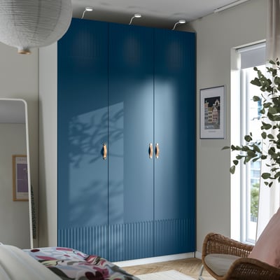 A large blue PAX wardrobe with gold handles, situated between a bed and a window with a plant.