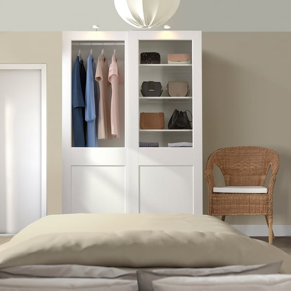 Modern bedroom with white PAX wardrobe showcasing clothes, beside wicker chair.