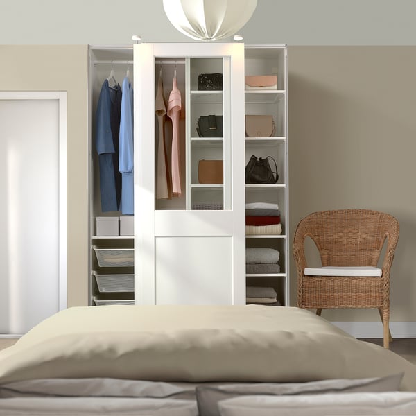 Bedroom scene with white PAX wardrobe, large bed, and wicker chair. Wardrobe, taller than the chair, holds clothes, bags, and folded items.