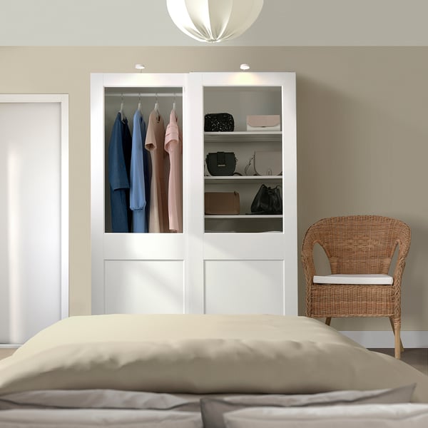 Modern bedroom with white PAX wardrobe, glass doors, and wicker chair beside bed with beige bedding.