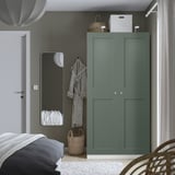 Minimalist bedroom with green PAX wardrobe, dark bedding, sheer curtains, and mirror. Dark green wardrobe contrasts with light grey walls.
