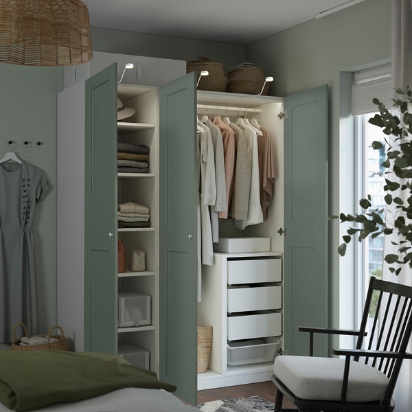A modern bedroom with a built-in wardrobe, featuring green doors, interior shelves, drawers, and clothes hangers. A chair and plant are placed nearby.