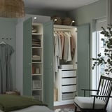 A modern bedroom with a built-in wardrobe, featuring green doors, interior shelves, drawers, and clothes hangers. A chair and plant are placed nearby.
