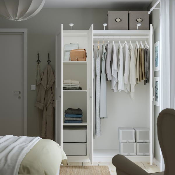 Modern bedroom with open white wardrobe showing clothes, folded clothes, and storage boxes, next to a bed with beige bedding.