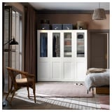 A modern, white PAX wardrobe with glass doors showcasing clothes, flanked by a wicker chair and a bed on a pink rug, with grey storage boxes atop.