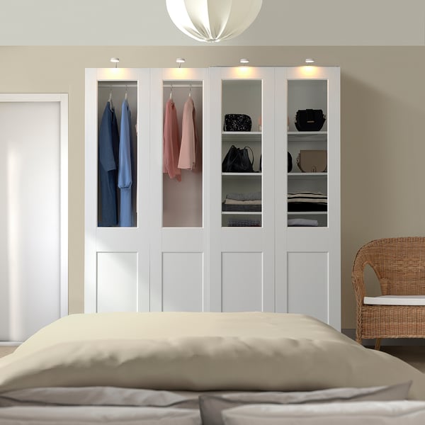 Neat bedroom with a white PAX wardrobe and glass doors, showing clothes. Near a large bed with beige bedding and a wicker chair.