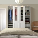 Minimalist bedroom with tall PAX wardrobe, glass & frosted panels, extensive storage. Complemented by beige pillows & wicker chair.