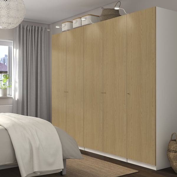 Large light-colored wooden closet with multiple doors next to a bed; white comforter and gray curtains visible.