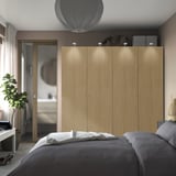 Modern bedroom with a large light wood PAX wardrobe, gray bed, desk with plant, and natural light.