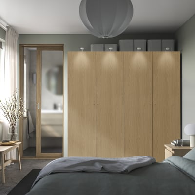 Modern wooden PAX wardrobe with soft-close doors and lighting. Two boxes on top, beside a door, next to a bed and vase.
