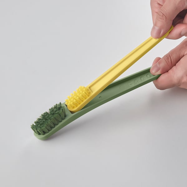 Hand holds yellow-green shoe brush tool. Top brush removes dirt, bottom scraper cleans edges. Bright colors. Designed for shoe cleaning.