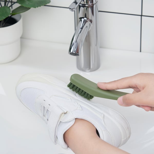 A hand cleans a white sneaker with a green brush by a sink.
