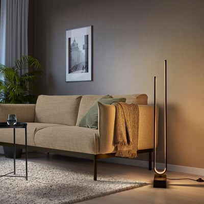 Modern living room with PILSKOTT floor lamp, beige sofa, and plant. Lamp has a sleek, tall, black design with warm lighting.