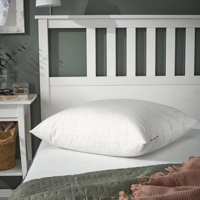 Bedroom scene featuring large white PILSPINNARE pillow on bed, accented with green and pink throws. Nightstand with lamp and decor left side.