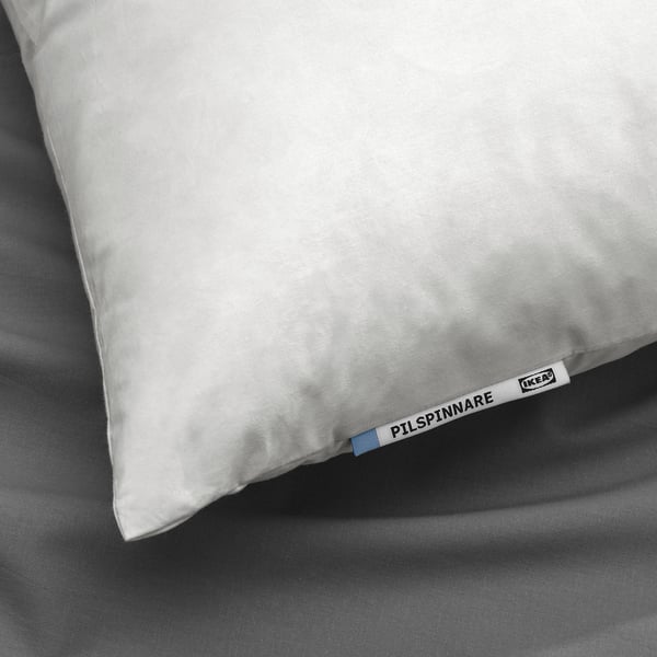 Image shows a single white pillow with ikea tag labeled PILSPINNARE. The pillow appears soft and plump, placed on a gray bedspread.