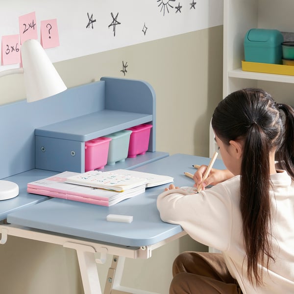 A girl sits at a PIPLÄRKA kids desk, tilting the surface for better posture. She writes in a workbook beside colorful storage bins and a lamp.