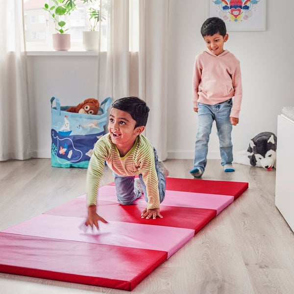 Kids playing indoors on colorful mat; one does somersaults, other jumps.
