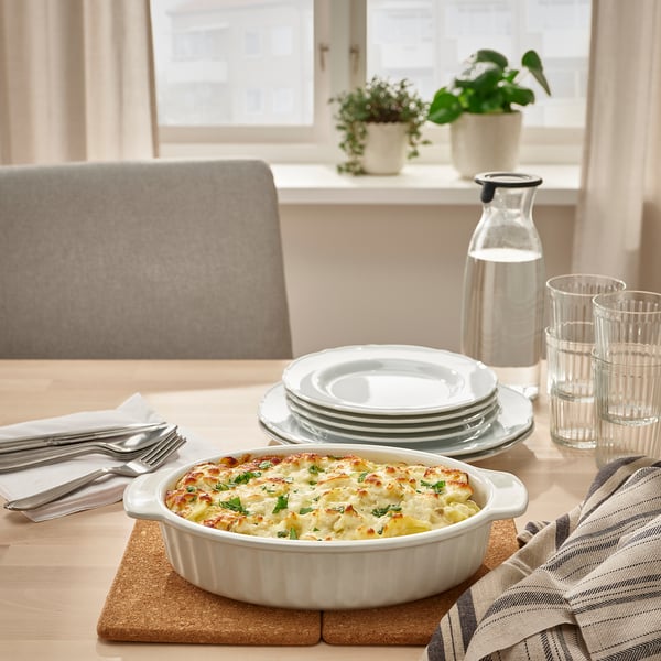 White stoneware baking dish with handles, filled with food, placed on wooden board on table near stacks of plates, set glasses, and silverware.