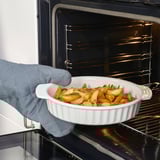 Hand holds white baking dish with roasted vegetables, placed inside oven. Dish has handles for easy lifting.