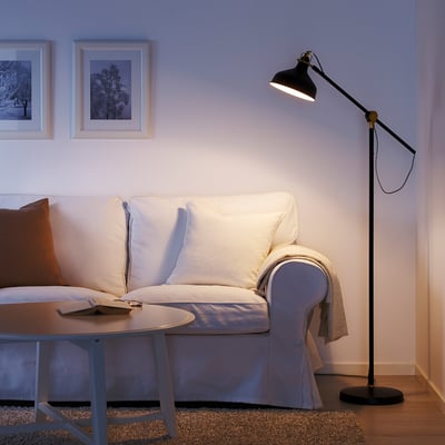 Cozy living room with white sofa, RANARP black floor lamp, brown rug, coffee table, and framed art. Lamp stands tall beside the couch.