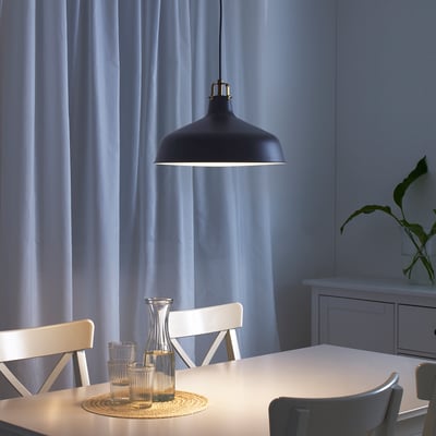 Modern dining area with black pendant lamp and white table, focused lighting on carafe and glasses, blue curtains, and white chairs.