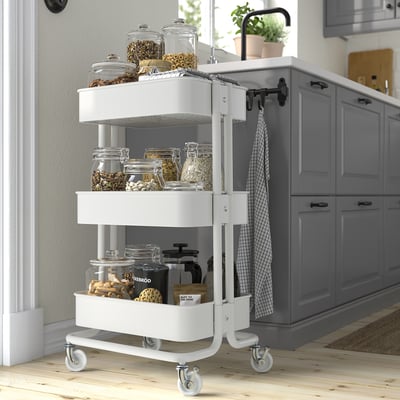 White ikea utility cart in kitchen, holding jars and supplies, next to cabinets.