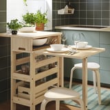 Modern kitchen setup with RESARÖ light wood table, two stools, plates, glasses, and herb pots on tiled wall backdrop.