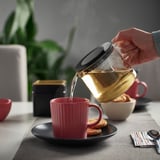 Person pours tea from a glass teapot with an infuser into a red mug, sitting on a black saucer next to cookies and a spoon.