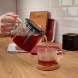 A hand pours tea from a glass RIKLIG teapot with an infuser into a clear mug on a wooden countertop.
