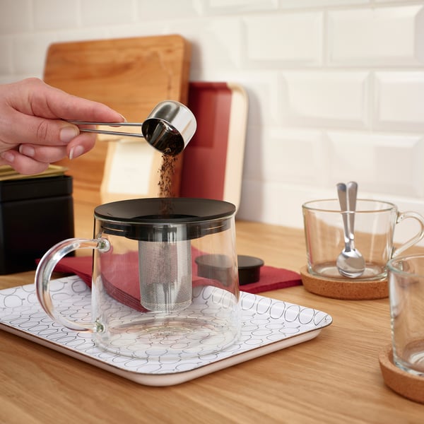 Person pours tea leaves into RIKLIG teapot with infuser on a tray.