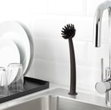 Black kitchen brush with suction cup base for scrubbing, resting on stainless steel sink near silver faucet.