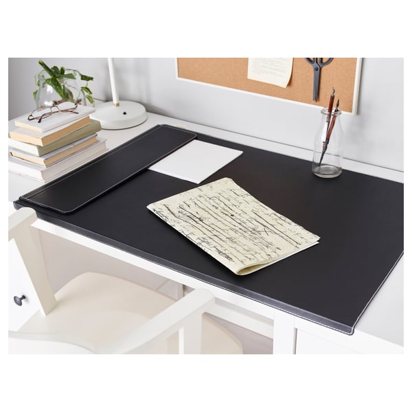 Black desk pad protects white desk, stays in place with weighted edge. Surrounded by books, lamp, pen vase, and cork board.