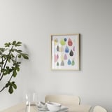A colorfully framed picture of various shapes hangs on a light wall above a wooden table with a white bowl and glass.