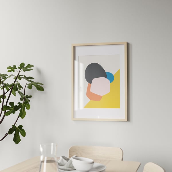 A modern wooden picture frame hangs on a wall, displaying colorful abstract art. Below it, a dining table holds glasses, cups, and a plant.