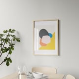 A modern wooden picture frame hangs on a wall, displaying colorful abstract art. Below it, a dining table holds glasses, cups, and a plant.