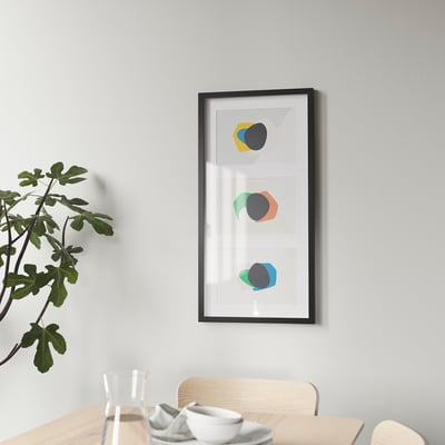 Modern wall art: abstract shapes in colorful frames, displayed vertically above a wooden table with a glass and bowl, accompanied by a leafy plant.