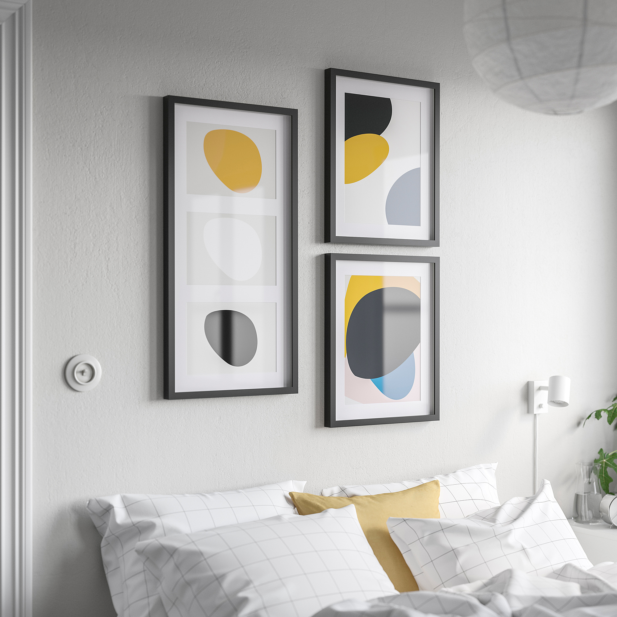 Three RÖDALM frames with bold art above bed, yellow & grey shapes, rectangular, modern, black, between lamp & door, bed has yellow checkered pillows.