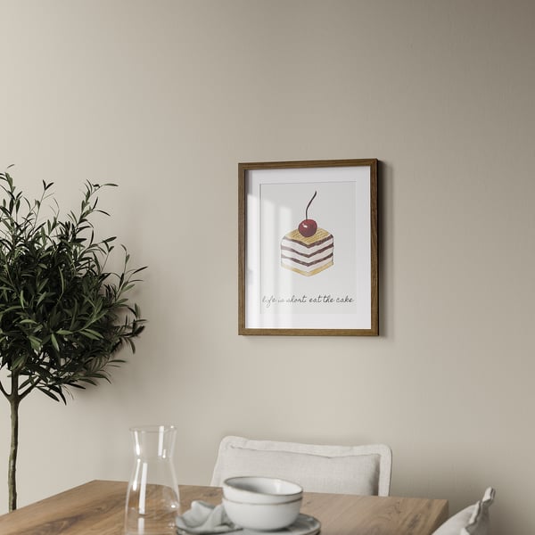 A framed picture featuring cake art hangs above a wooden table with a cup, saucer, and carafe.