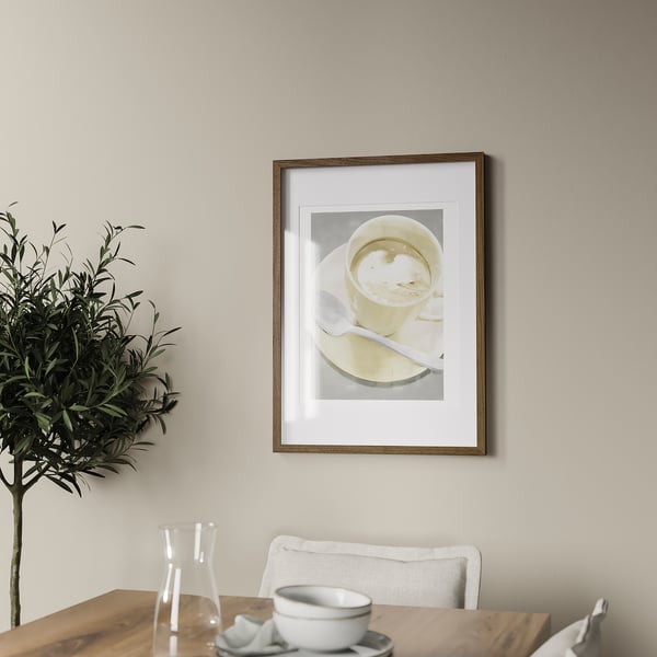 A coffee cup hangs on a wall above a wooden table with a white chair. A plant and carafe are on the table. The frame holds a picture up to 50x70 cm.