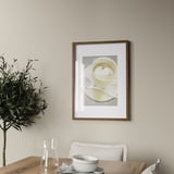A coffee cup hangs on a wall above a wooden table with a white chair. A plant and carafe are on the table. The frame holds a picture up to 50x70 cm.