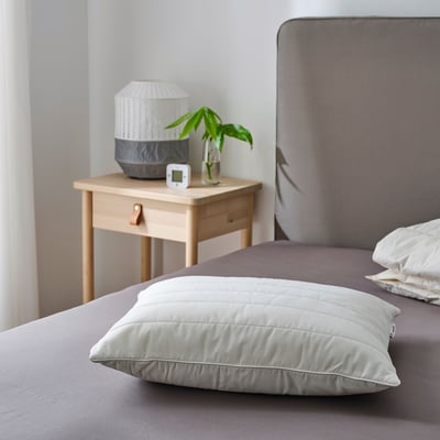 Bedroom with grey RUMSMALVA pillow on purple bed, wooden side table with lamp and plant.